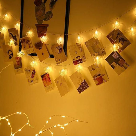 Heart shaped LED string lights emitting warm white light decorating a wall with hanging photos for cozy home ambiance
